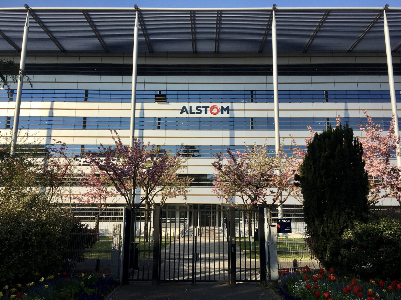 A modern office building with "Alstom" logo, featuring large glass windows and surrounded by flowering trees and shrubbery, secured by a metal fence at the foreground.