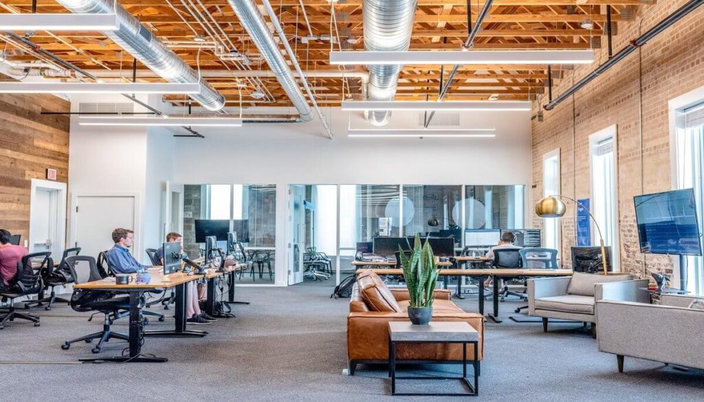 Spacious modern office with high ceilings, exposed ductwork, wooden accents, and several people working at desks. A sofa and plant are in the foreground. Natural light comes through large windows, while innovative commercial parking solutions like Wayleadr optimize the external parking lot systems.