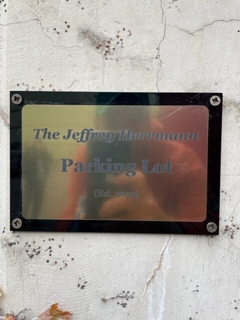 Plaque on a wall reads: "The Jeffrey Herrmann Parking Lot (Est. 2022) – Providing convenient visitor parking and serving as a prime spot for commercial parking.