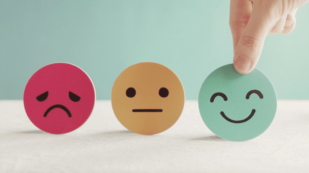 Three circles with faces: a red sad face, a yellow neutral face, and a green happy face. A hand is picking up the green happy face.