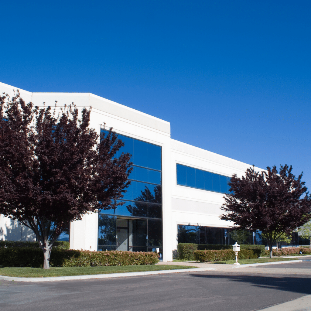 A modern, two-story office building with large blue-tinted windows, white exterior walls, and landscaping that includes trees and shrubs on a clear day features sophisticated parking management powered by Wayleadr.