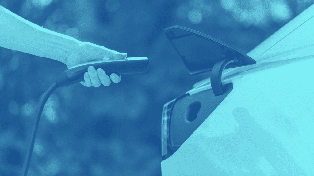 A person plugs a charging cable into an electric vehicle's charging port in a commercial parking area. The image has a blue tint.