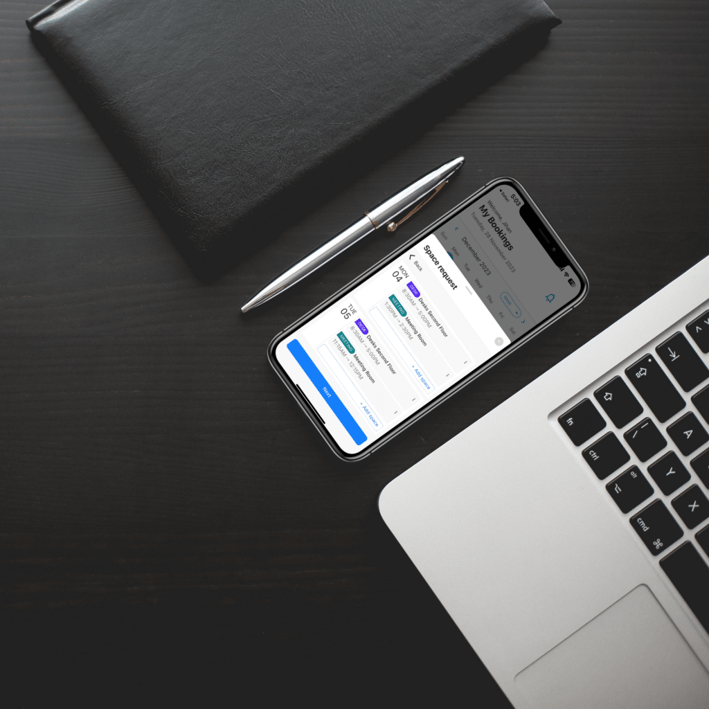 A smartphone displaying calendar events next to an open laptop, a pen, and a closed black notebook on a dark wooden desk—perfect for managing your day, including meetings about visitor parking solutions.
