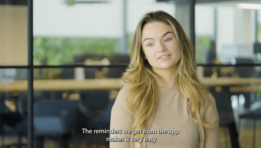 A person with long, light brown hair wearing a beige top is speaking. The caption reads, "The reminders we get from the app makes it very easy." The background shows a modern office space with visible visitor parking signs in the distance.