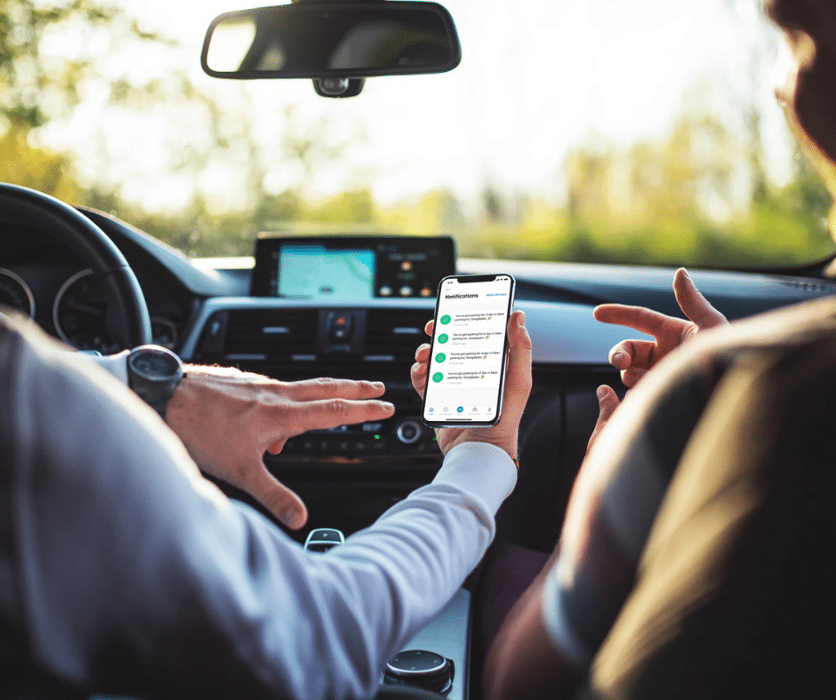 Two people are in a car, one holding a smartphone showing a messaging app, while the other points at the screen. The car's GPS navigation system is visible in the background, suggesting they might be navigating to a complex with advanced parking lot systems.