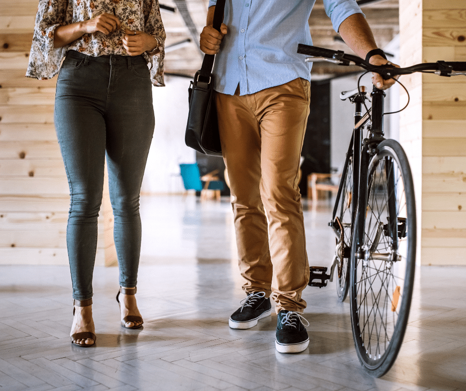 Two people stand indoors, one holding a bicycle by its handlebars. Both wear casual clothes and their faces are not visible. In the background, posters advocate for efficient parking lot management through Wayleadr's commercial parking solutions.