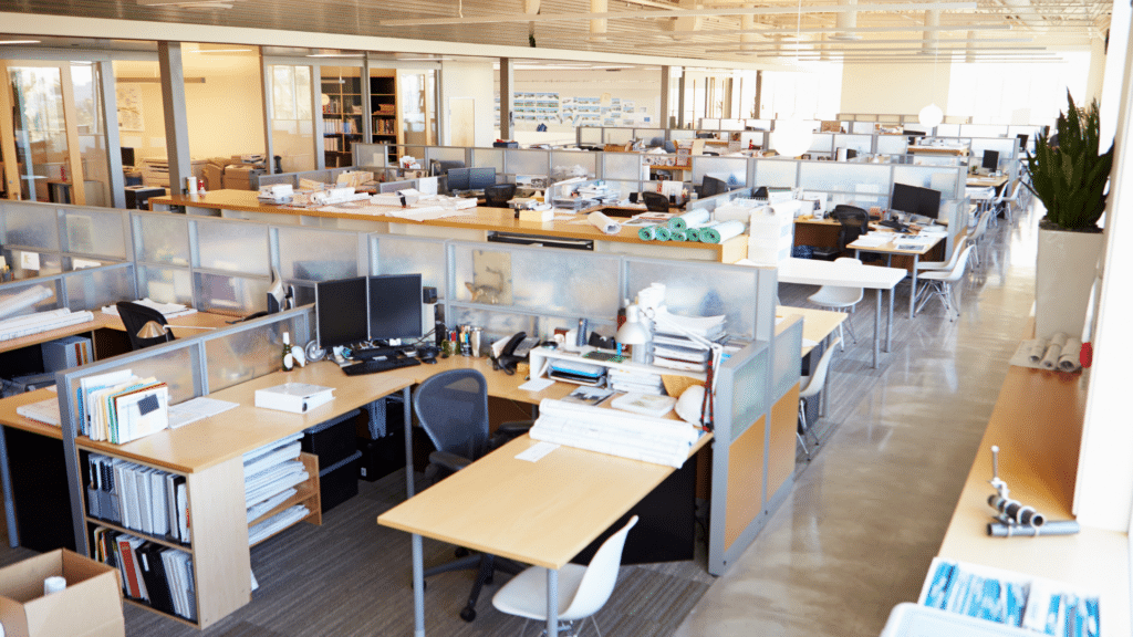A spacious open-plan office with rows of workstations, each equipped with computers, office supplies, and personal items. The room is brightly lit with natural light from large windows and offers a clear view of the visitor parking area, ensuring smooth parking lot management.