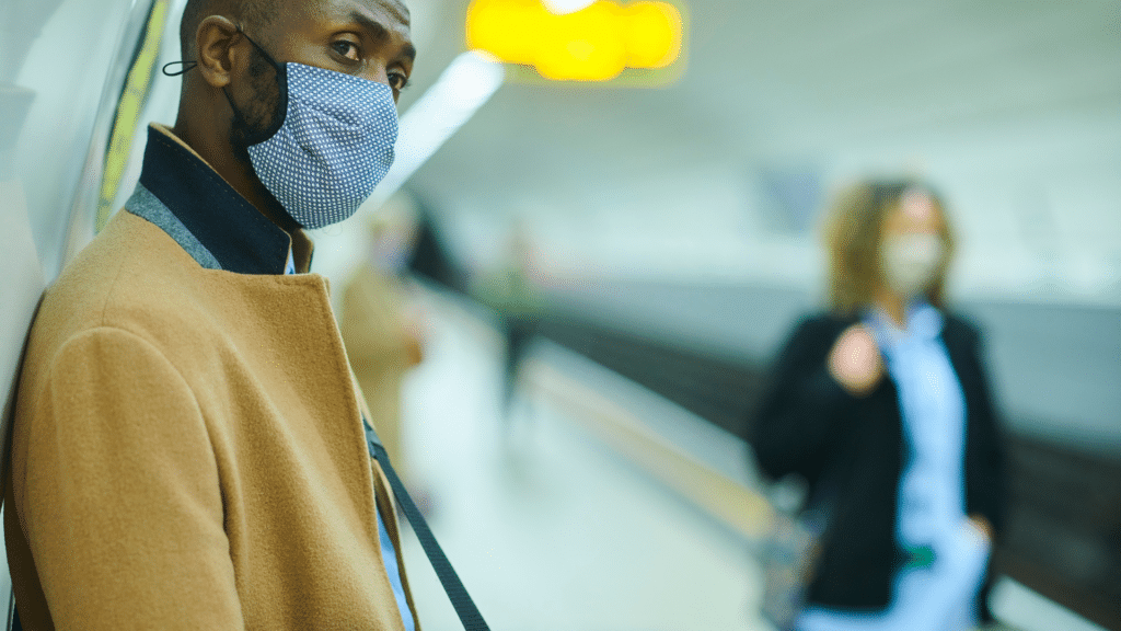 A man wearing a coat and face mask leans against a subway wall, while blurred commuters in the background, also masked, hurry by. He's perhaps contemplating wayleadr's innovative parking management solutions for visitor parking in his busy city.