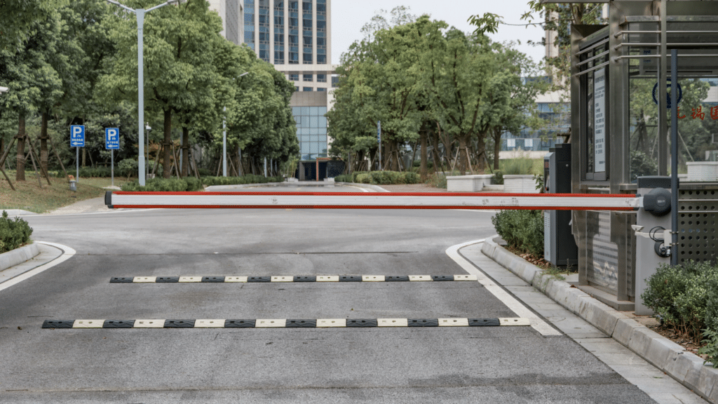 A closed boom gate blocks a road near a commercial parking area. A pedestrian crossing is visible in front of the gate. Trees line the road, and a tall building is seen in the background, likely managed through advanced parking lot systems like Wayleadr.