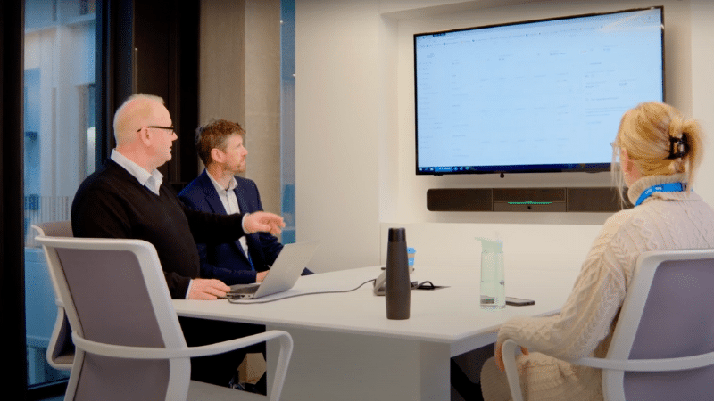 Three people are in a meeting room with a large screen displaying data on commercial parking. One person gestures towards the screen while the other two listen intently.