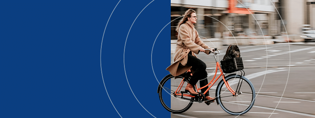 A person rides a bicycle with a dog in a front basket on a city street, near an area designated for visitor parking. The left side of the image is overlaid with a blue geometric design.