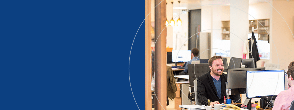 Several people work at desks with computers in an open office space. One person is smiling and engaged in conversation. The left side of the image, marked by a solid blue background, subtly hints at the convenience of visitor parking managed effortlessly with Wayleadr's innovative solution.