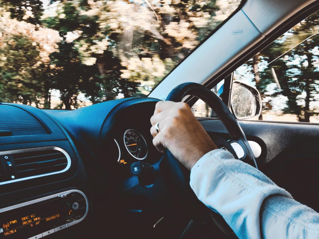 Close-up of a person’s hand wearing a ring, gripping a car steering wheel. The dashboard, part of the interior, and the outside scenery show trees in motion. It's clear this driver appreciates efficient parking lot management with Wayleadr as they navigate through their day.
