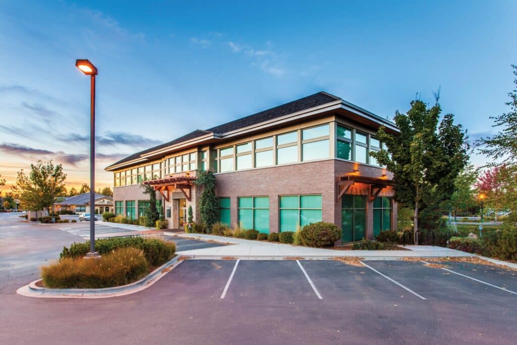 A modern two-story office building with large windows, surrounded by landscaped greenery and an adjacent parking lot, employs Wayleadr for efficient parking lot systems, pictured at dusk.