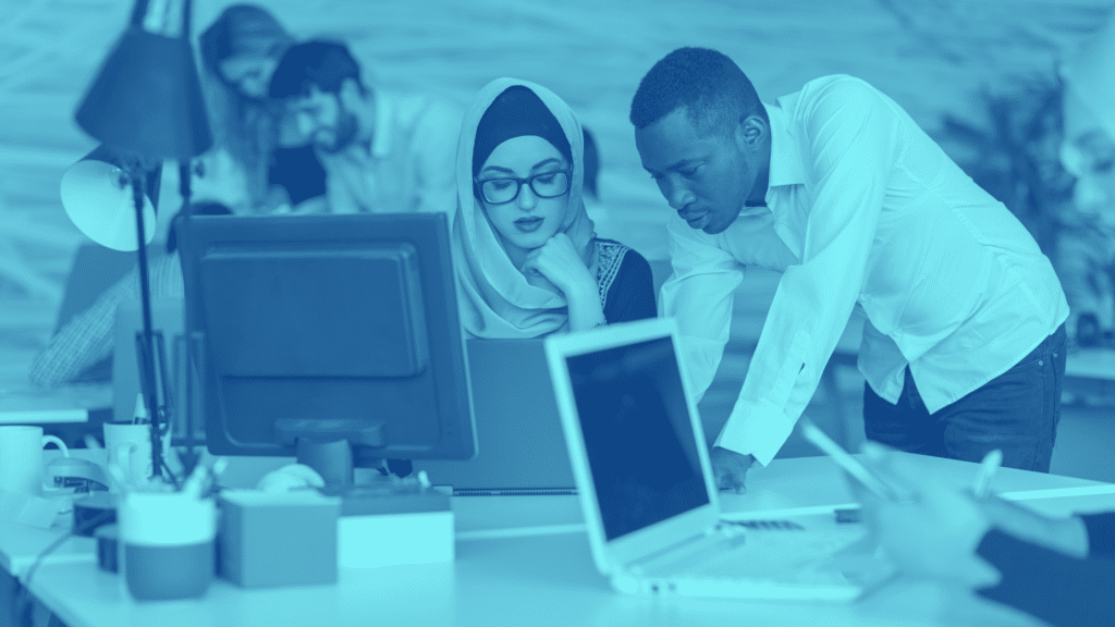 A group of people working together in an office, with two individuals focused on a computer screen in the foreground, possibly discussing parking lot management.