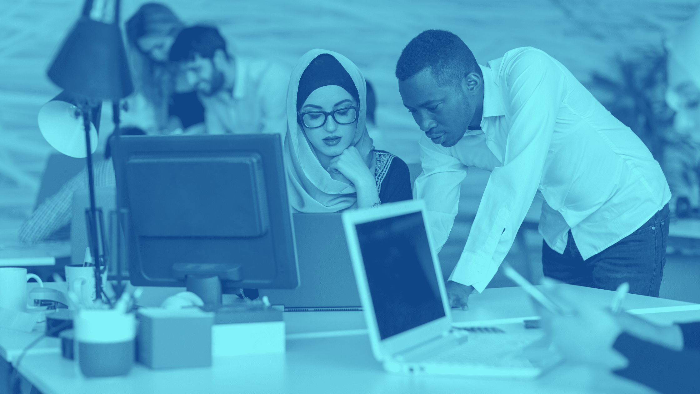 A group of people working together in an office, with two individuals focused on a computer screen in the foreground, possibly discussing parking lot management.