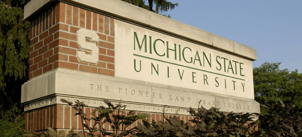 Sign for Michigan State University made of brick and stone, featuring the university's name in green text and the letter "S" on the left side. The sign includes the subtitle "The Pioneer Land Grant College," located near visitor parking for easy access.