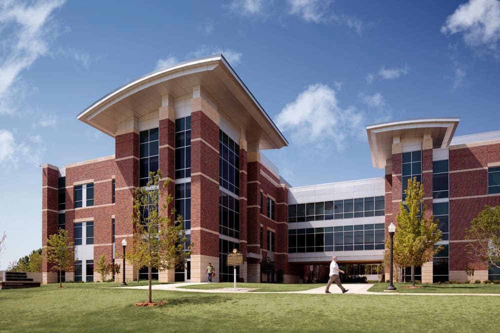 Exterior view of a modern three-story brick and glass building with landscaped surroundings. Two people walk on the grass in front of the building under a sunny sky, appreciating its visitor parking managed efficiently by Wayleadr.