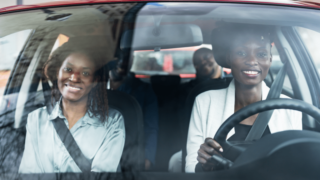A woman driving a car with three other passengers, all smiling and wearing seatbelts, navigates through the commercial parking lot.
