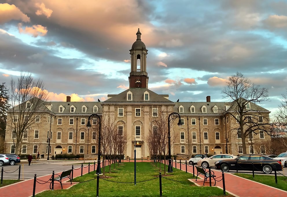A large, historic building with a clock tower at the center, surrounded by a landscaped walkway and trees. The sky features a mix of clouds and colorful sunset hues, while parking lot systems ensure organized parking for the nearby cars.