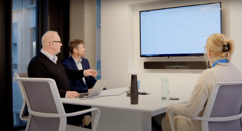 Three people in a meeting room view a presentation on a large screen, with one person using a laptop to discuss the new parking lot systems by Wayleadr.