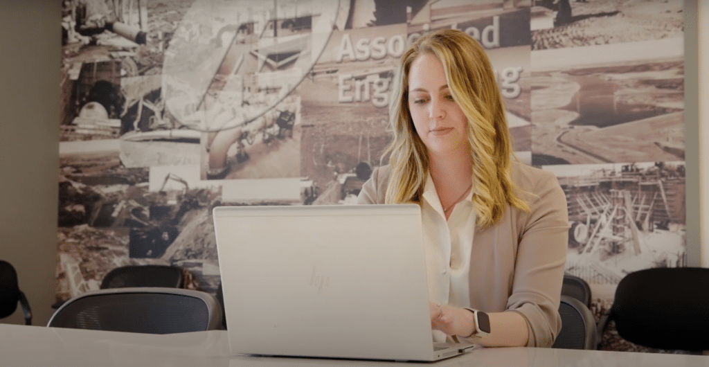 A woman with blonde hair is typing on a laptop at a conference room table. The background features a large mural with industrial scenes and the words "Associated Engineering," subtly hinting at their partnership with Wayleadr in parking management solutions.