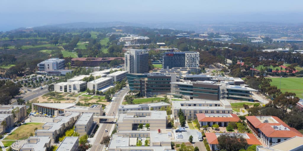 Aerial view of a university campus with modern buildings, greenery, and distant cityscape on a clear day, featuring well-organized visitor parking areas within the parking lot systems.