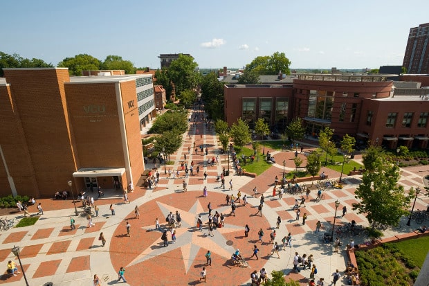 Aerial view of a college campus with brick buildings, walkways, trees, and students walking and gathering around a large star design in the central courtyard. Visitor parking is conveniently located nearby for efficient parking management.