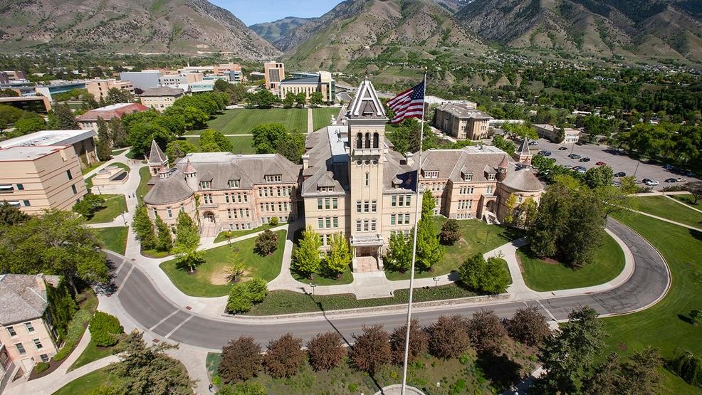 Aerial view of a historic university building with a clock tower, surrounded by trees and mountains. An American flag flies prominently in the foreground, near the visitor parking area nestled among the picturesque landscape.