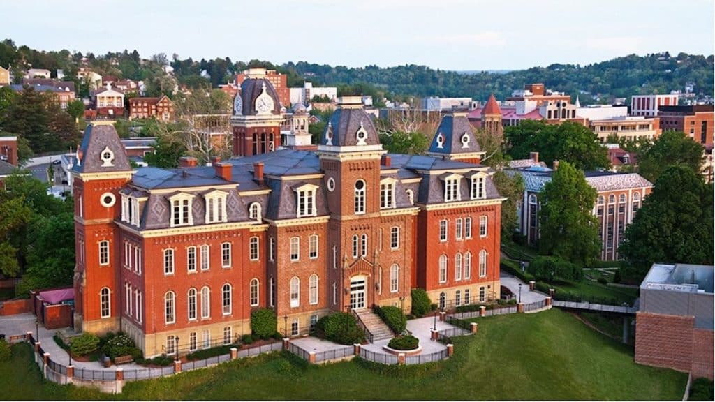 A large red-brick university building with multiple towers and a green lawn is surrounded by trees, visitor parking, and other buildings in a hilly area.