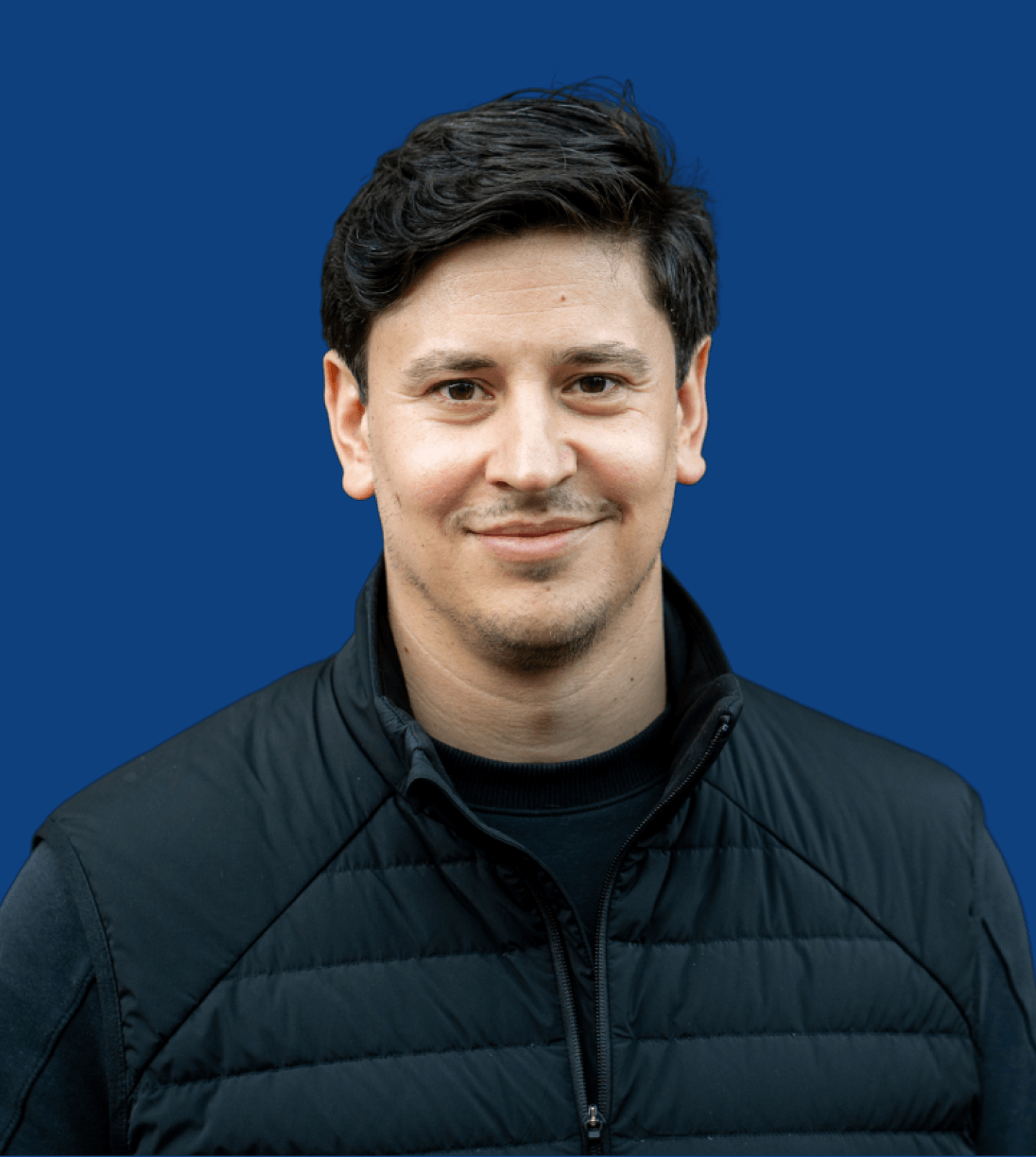 A man with short dark hair, wearing a black quilted jacket over a black shirt, stands in front of a solid blue background, smiling slightly.