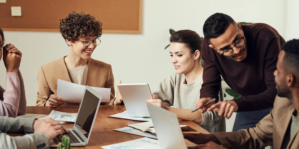 Five people sit around a table with laptops and documents, collaborating and discussing work in a modern office setting.