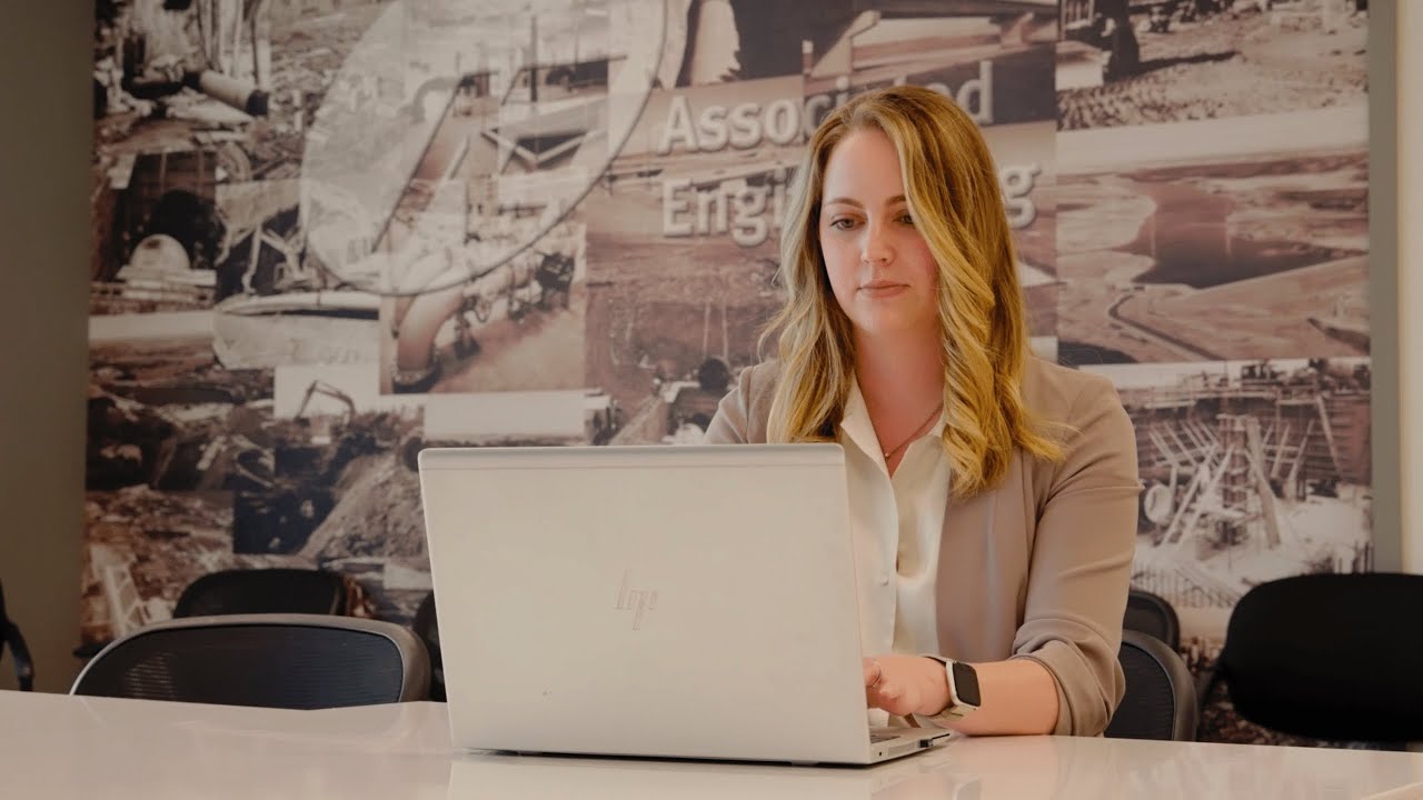 A woman with blonde hair sits at a conference table, working on a laptop. There is a mural with engineering images on the wall behind her.