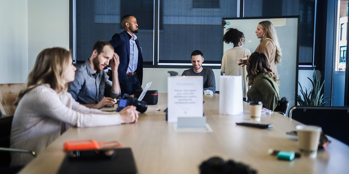 A group of people are gathered around a conference table in an office, some seated and some standing, engaged in discussion and using laptops and notebooks.