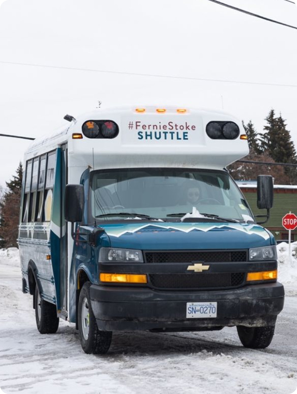 A blue and white shuttle bus labeled "#FernieStoke SHUTTLE" is parked on a snowy street near a stop sign.