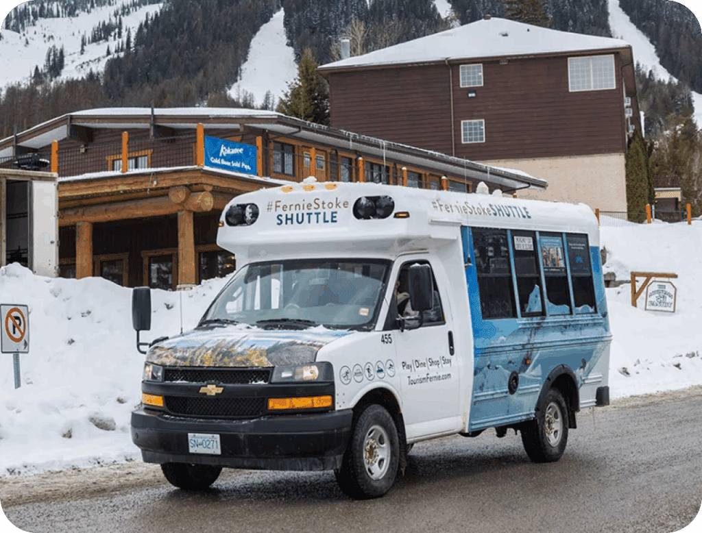 A white shuttle bus labeled “FernieStoke Shuttle” is parked on a snowy road in front of a lodge-style building, with snow-covered trees and mountains in the background.