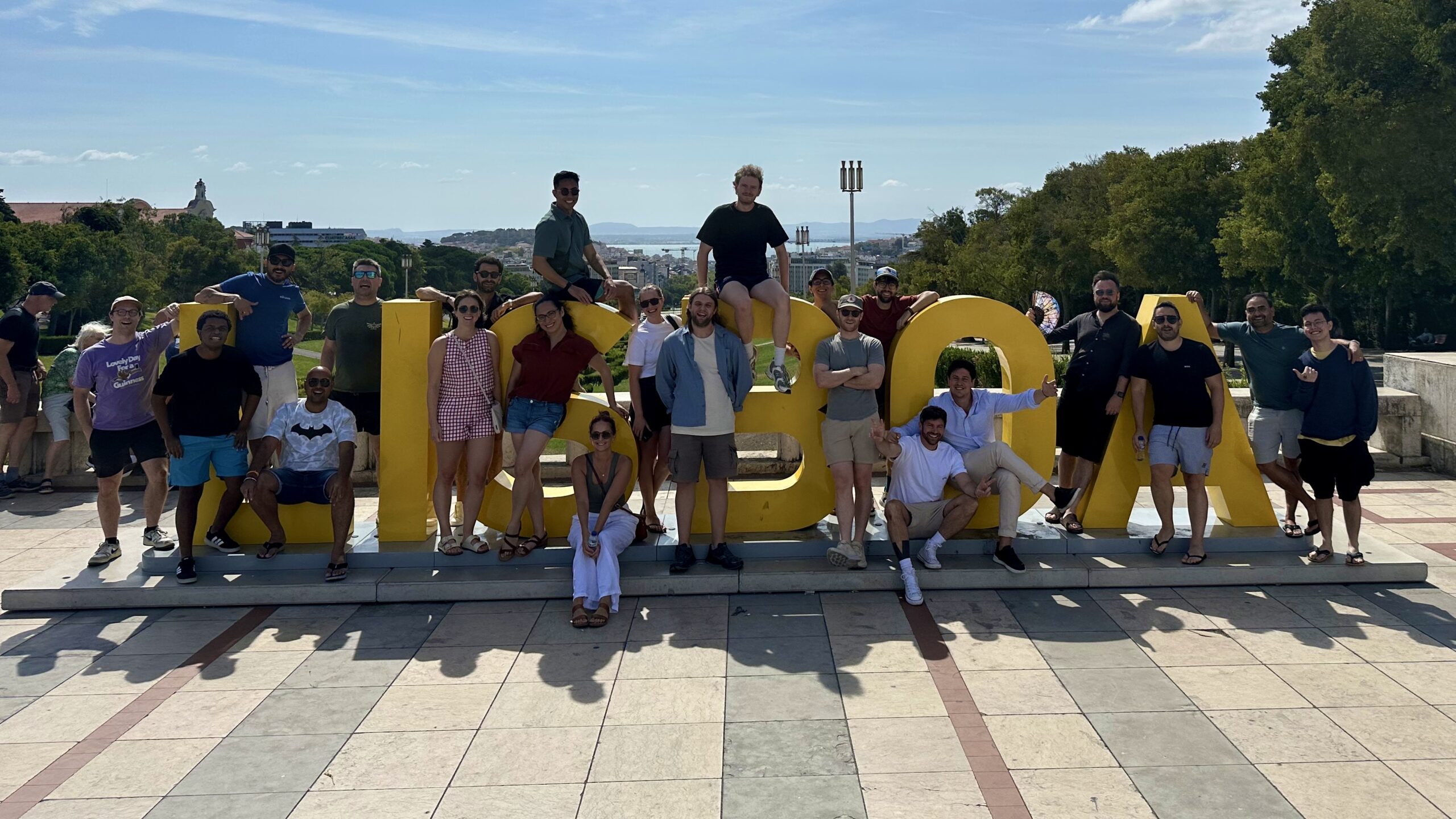 A group of people pose together around and on top of large yellow "LISBOA" sign outdoors on a sunny day.