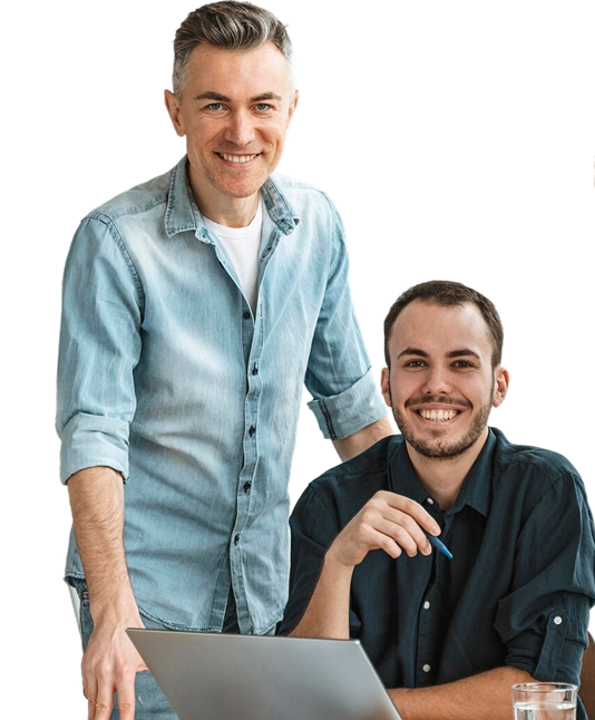 Two men pose together, one standing and one seated in front of a laptop, both smiling at the camera against a plain background.